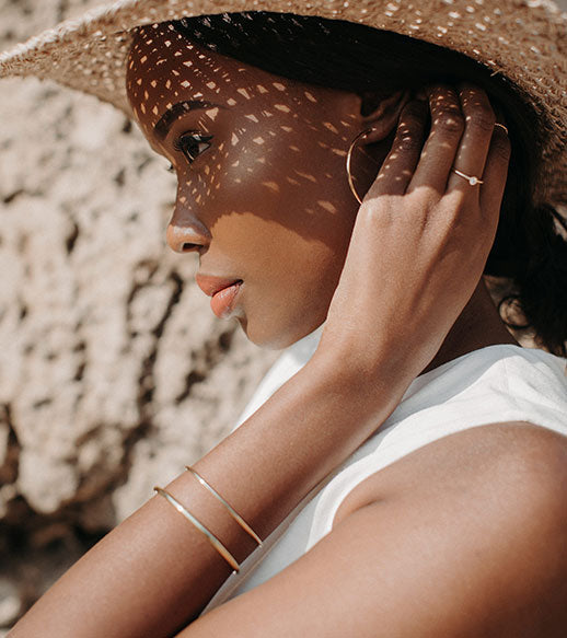 Woman wearing a wide-brimmed hat and minimalist 9ct gold oval cuff bracelets, styled with delicate rings, photographed in natural light.