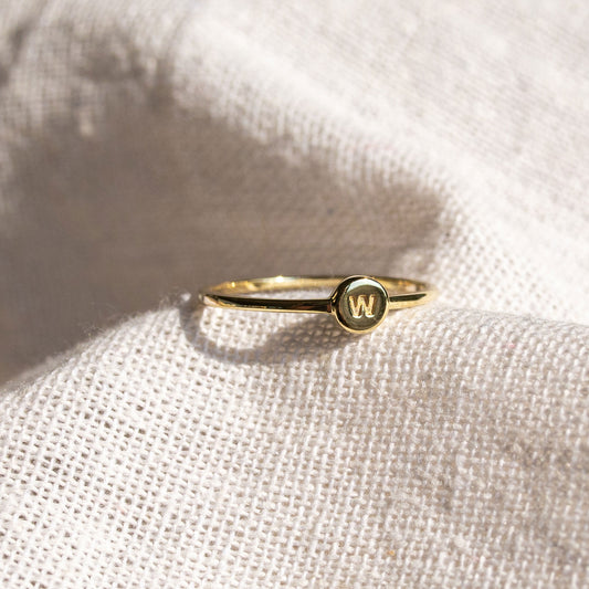 Close-up of a delicate 9ct gold signet stacker ring with an initial disc, photographed on textured fabric.