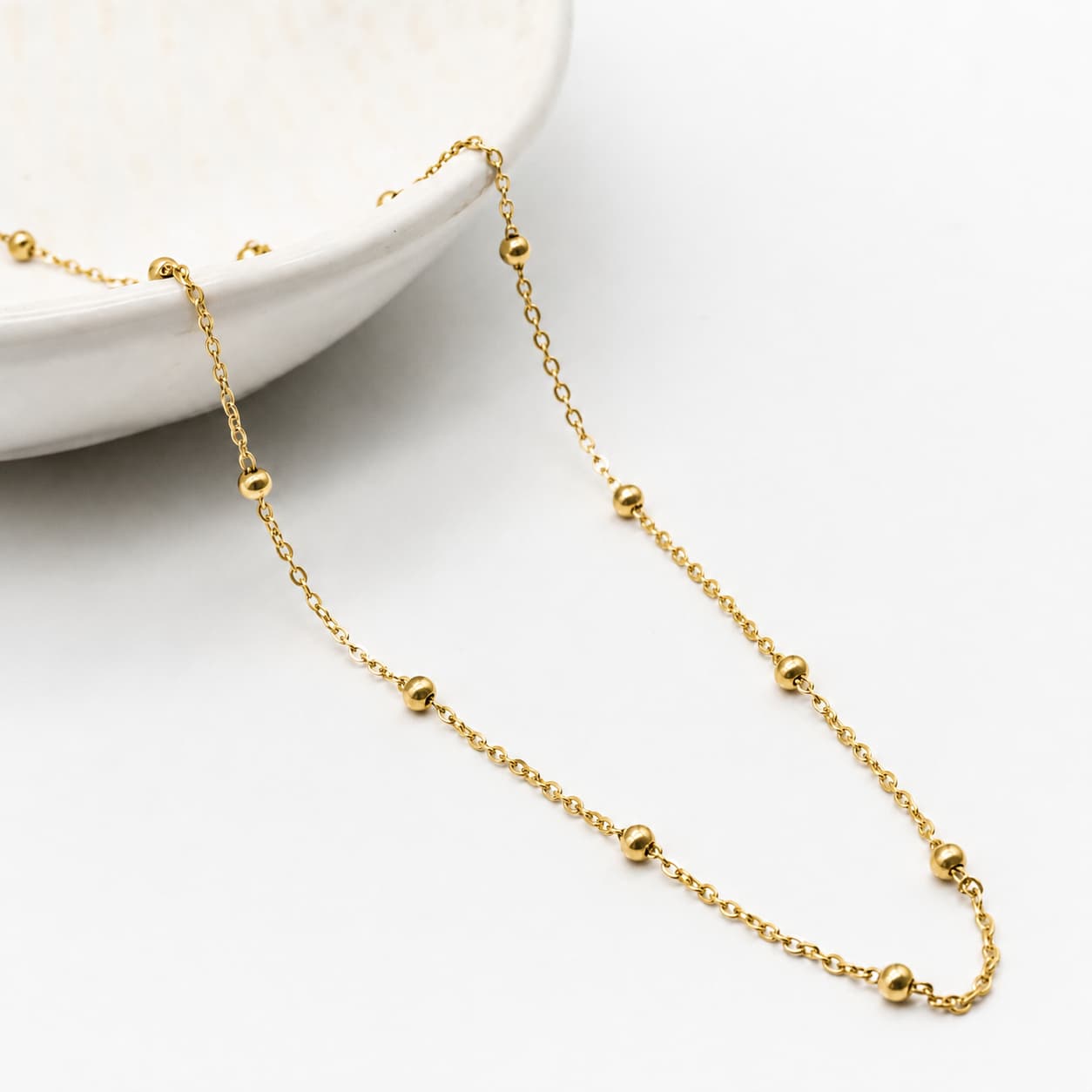 A delicate gold chain necklace with evenly spaced small gold beads, draped elegantly across a clean white surface next to a white ceramic dish.