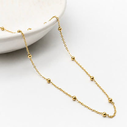 A delicate gold chain necklace with evenly spaced small gold beads, draped elegantly across a clean white surface next to a white ceramic dish.