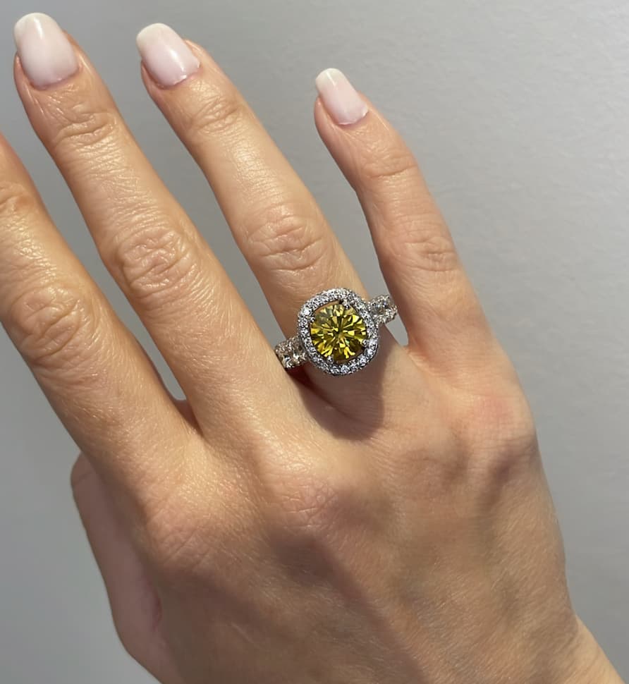 A close-up lifestyle view of a woman's hand wearing a luxury halo engagement ring featuring a large cushion-cut canary yellow diamond center stone, a brilliant diamond halo, and a split-shank pave diamond band
