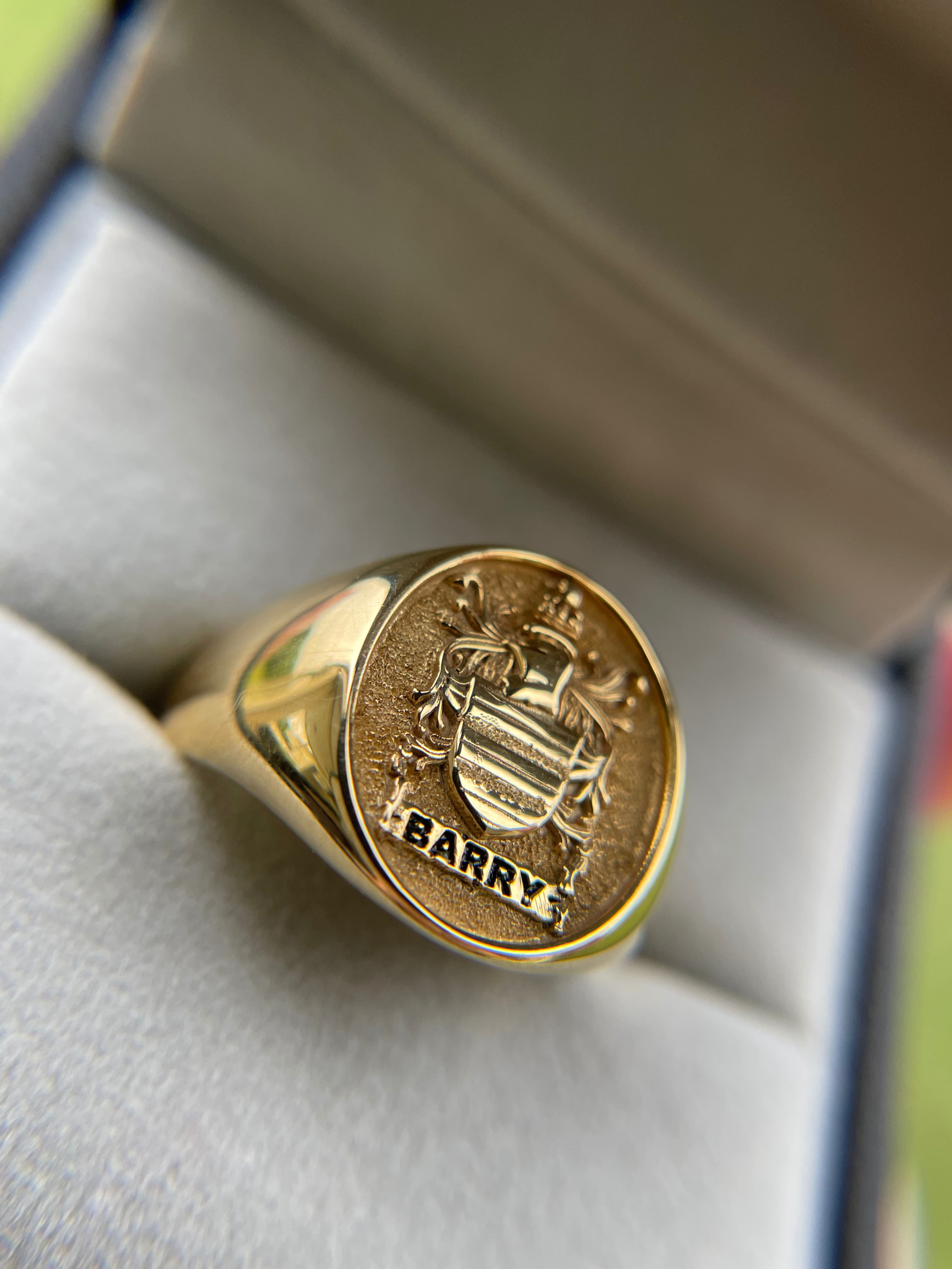 Close-up view of a heavy solid yellow gold signet ring featuring a custom family coat of arms crest and the name BARRY intricately engraved on the oval face, resting inside a luxury jewelry box.
