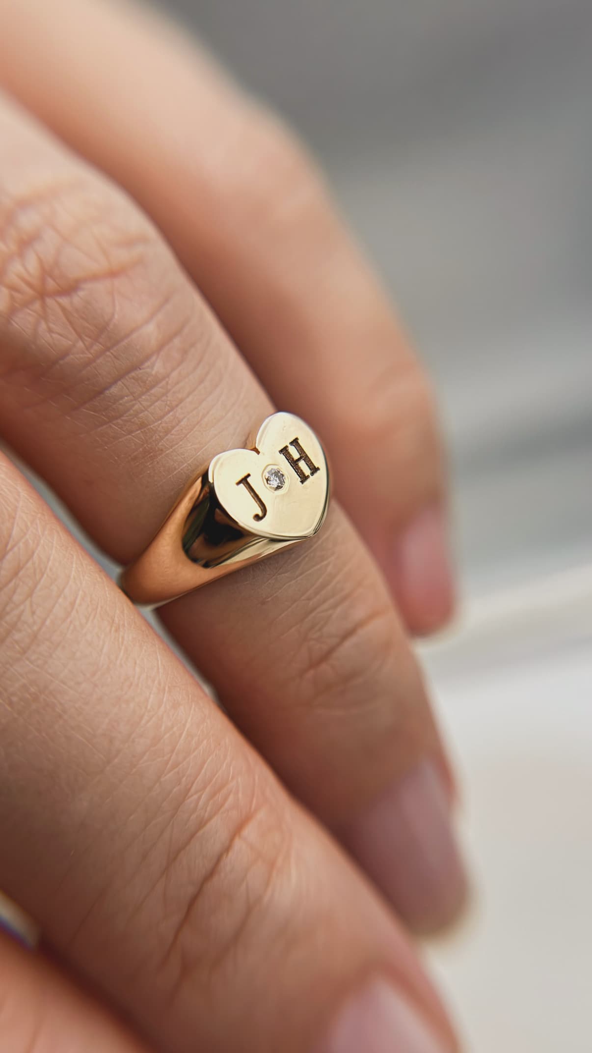 Close-up lifestyle view of a polished yellow gold heart-shaped signet ring engraved with the initials J and H, featuring a small round diamond starburst setting between the letters, worn on a pinky finger.