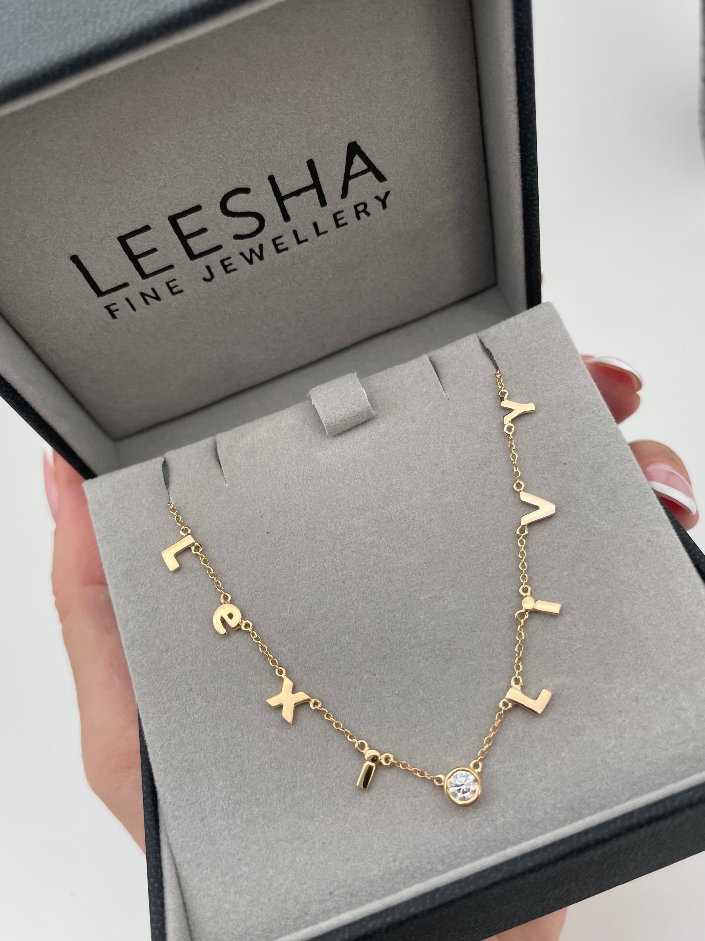 Personalised gold letter charm necklace featuring individual hanging initial letters and a small round bezel-set diamond charm, presented in a luxury branded LEESHA jewellery box.