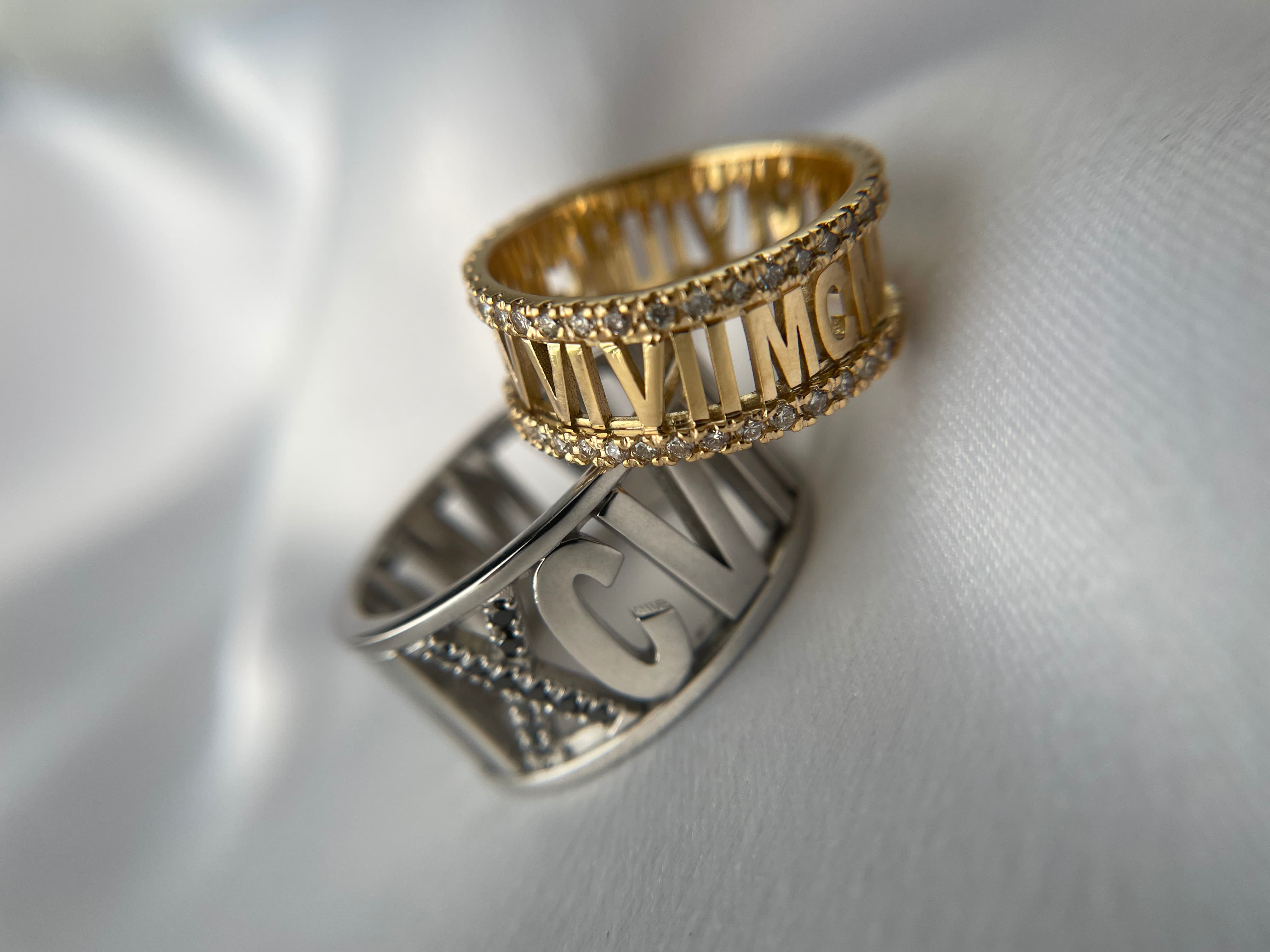 Two wide custom cutout band rings—one yellow gold ring featuring Roman numerals bordered by pave diamonds, and one white gold or platinum ring with bold block initials accented by black diamonds—displayed on white satin.