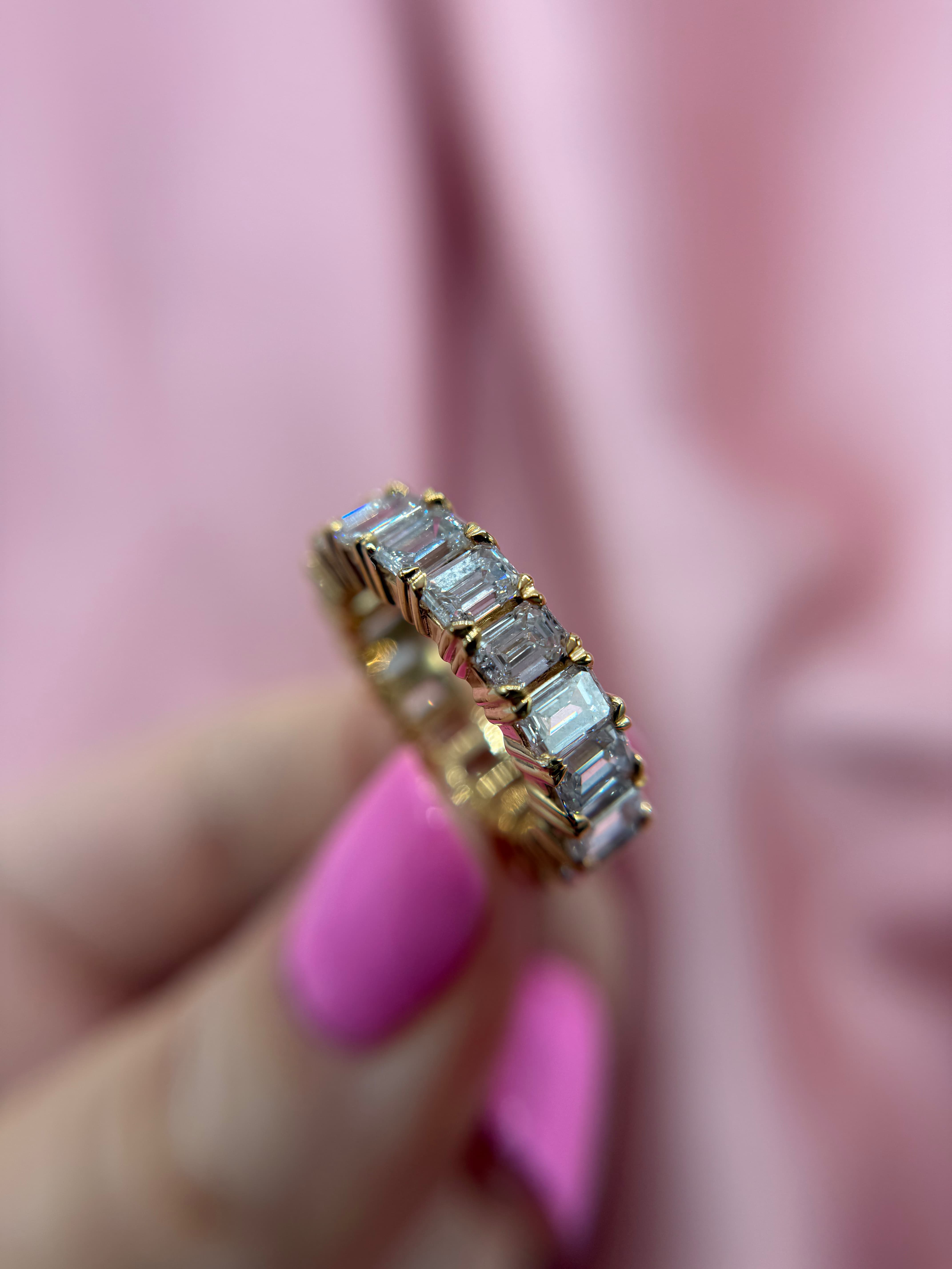 Close-up view of a luxury yellow gold eternity band ring set with a continuous row of brilliant emerald-cut diamonds, held by a hand with pink nail polish against a soft pink fabric background.