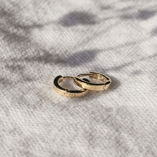 Close-up of polished yellow gold mini huggie earrings displayed on textured fabric.