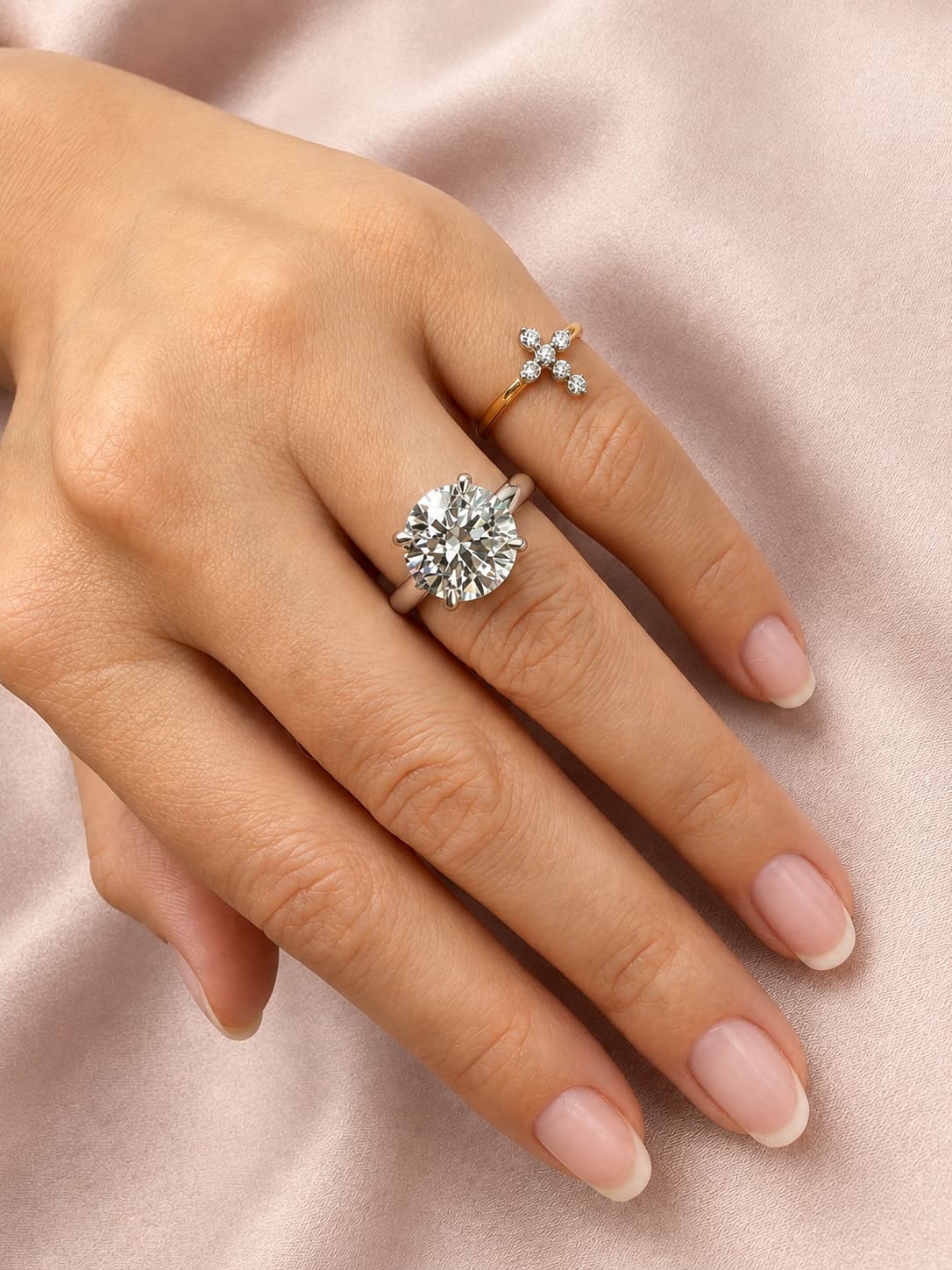 Close-up lifestyle view of a woman's hand wearing a large round brilliant-cut diamond solitaire engagement ring on her middle finger and a delicate yellow gold diamond cross ring, set against a soft pink satin background.