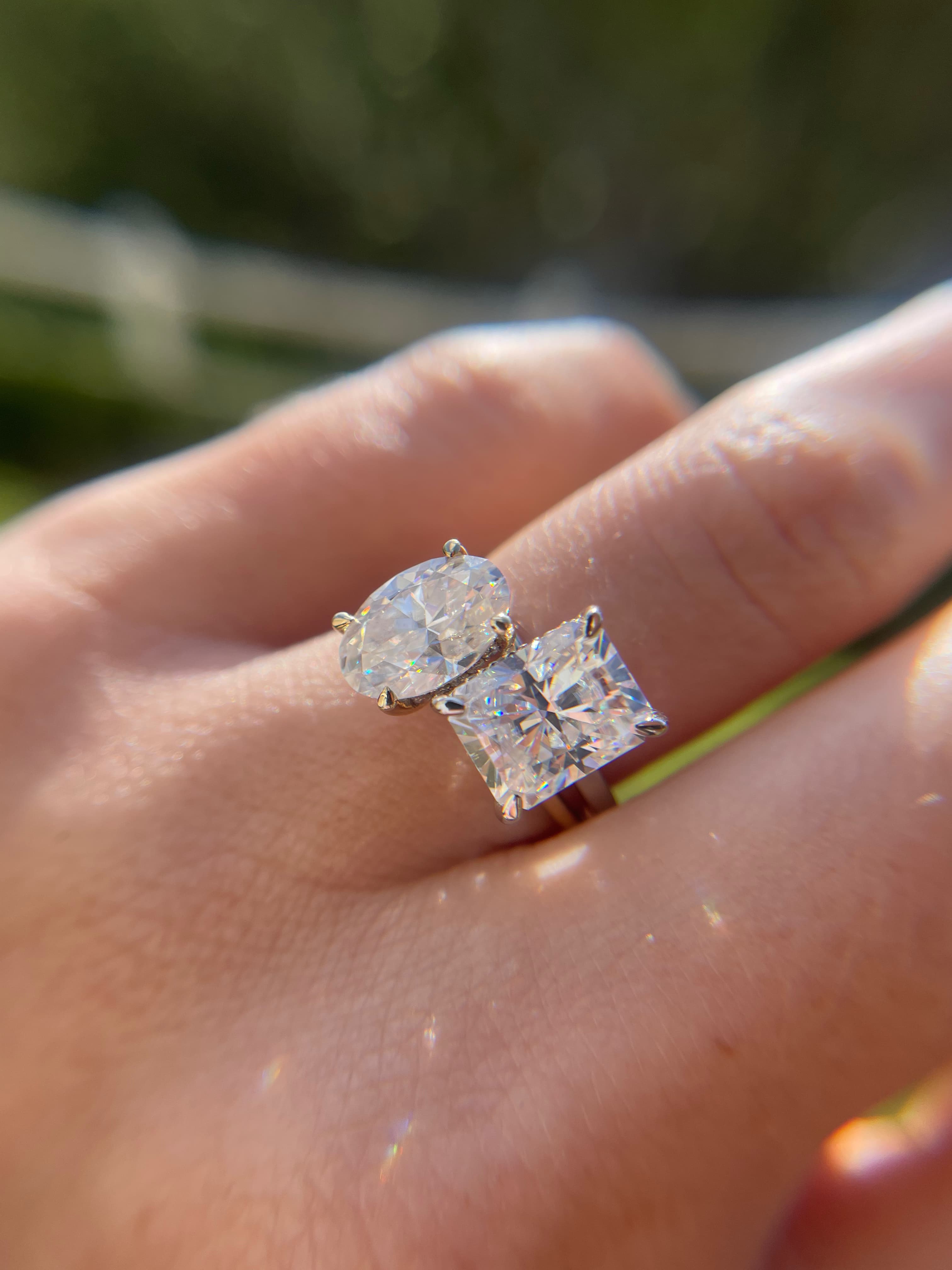 Close-up lifestyle view of a platinum Toi et Moi engagement ring featuring a brilliant oval-cut diamond paired with a radiant-cut diamond on a woman's hand in natural outdoor sunlight.
