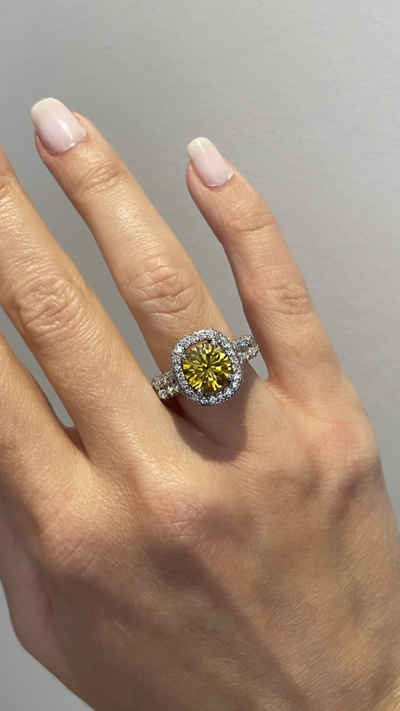 A close-up of a hand wearing a ring with a round yellow center stone surrounded by a halo and a silver-toned band, set against a plain light gray background.